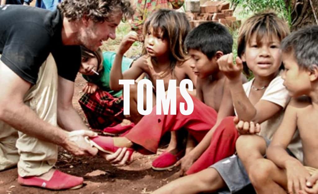 The text 'Toms' over the image of a man putting shoes on less privileged kids
