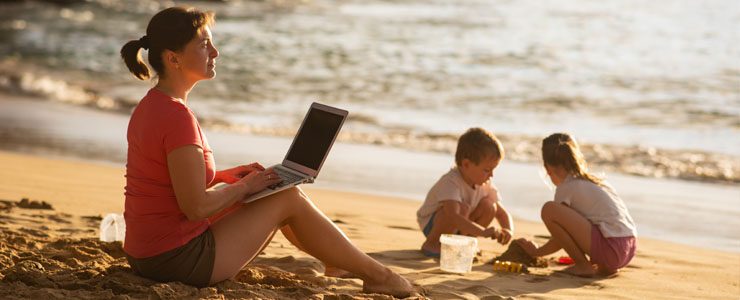 Woman on a beach working on a laptop while her children are playing